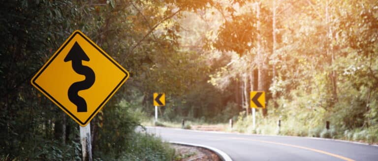 Focus is on a yellow caution sign showing there is a bend in the road on an unsafe road.