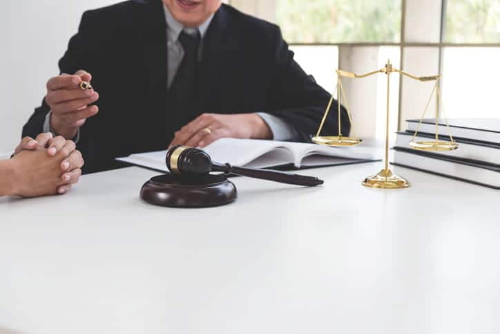 A dog bite attorney speaks with his client at his desk. In front of him is paperwork, a gavel, a stack of books and the scales of justice.