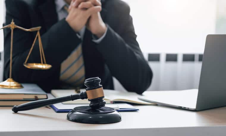 A medical malpractice lawyer is working at his desk. In front of him is an open laptop, paperwork, the scales of justice, and a gavel.