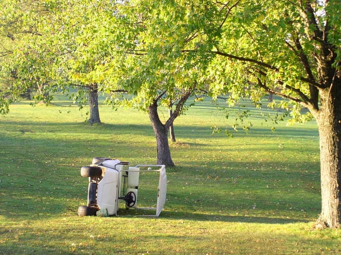 A golf cart tipped on its side under trees at a golf course.