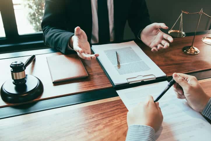 A personal injury attorney discusses a contract with his client. On his desk is a gavel, a notebook, paperwork, and the scales of justice.