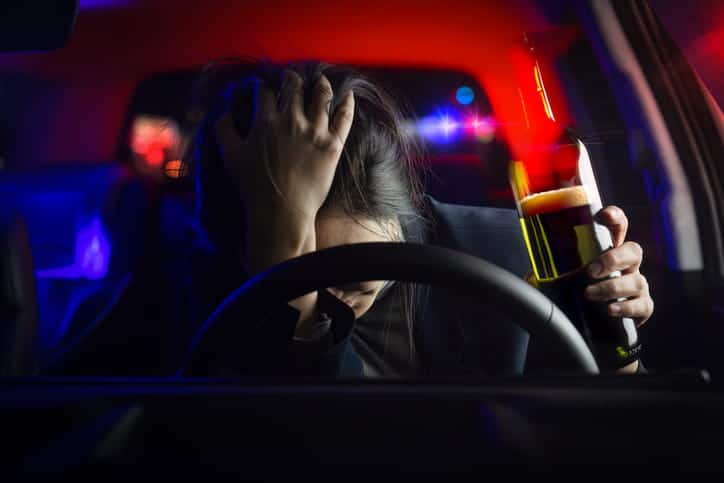 A woman holding a beer behind the wheel of a car with police lights behind her. Her head is in her hand as she leans over the steering wheel in distress.