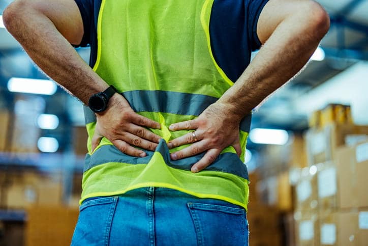 A worker in a safety vest is clutching his back in pain after a work injury.