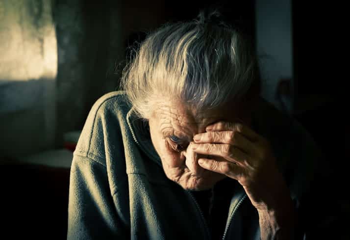 An elderly woman has a hand over her face as she sits in the dark.