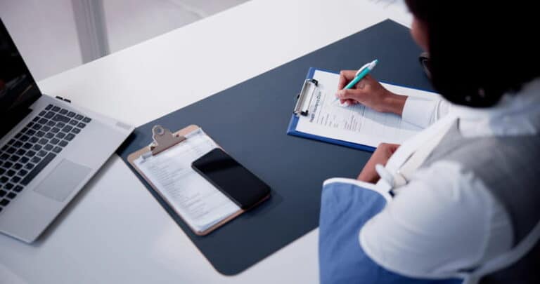 A woman in an arm sling is filling out health insurance forms with her phone, more paperwork, and laptop in front of her after being in a personal injury accident.