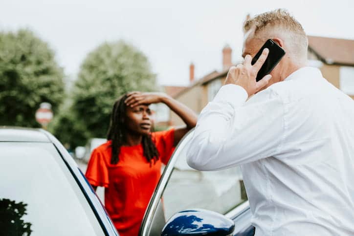 A man on the phone as a woman holds her head after they were in a car accident.