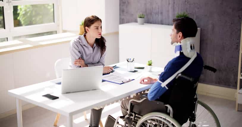 A man in a neck brace, arm sling, and sitting in a wheelchair speaks with his personal injury attorney.