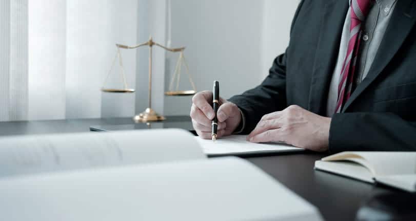 A personal injury attorney is writing down notes about a case at his desk. Next to him is the scales of justice.