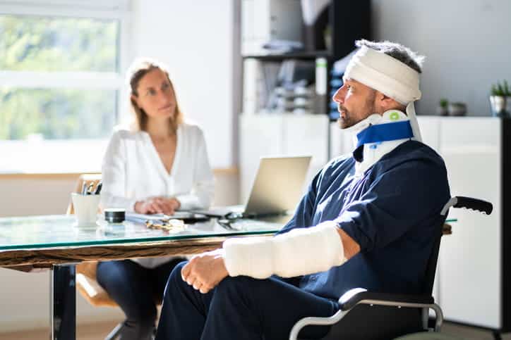 A man with his head and arm wrapped while wearing a neck brace sits in a wheelchair as his personal injury attorney discusses his case.