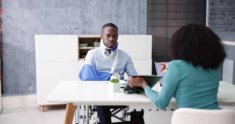 A man in a neck brace, arm sling, and sitting in a wheelchair meets with his personal injury attorney.